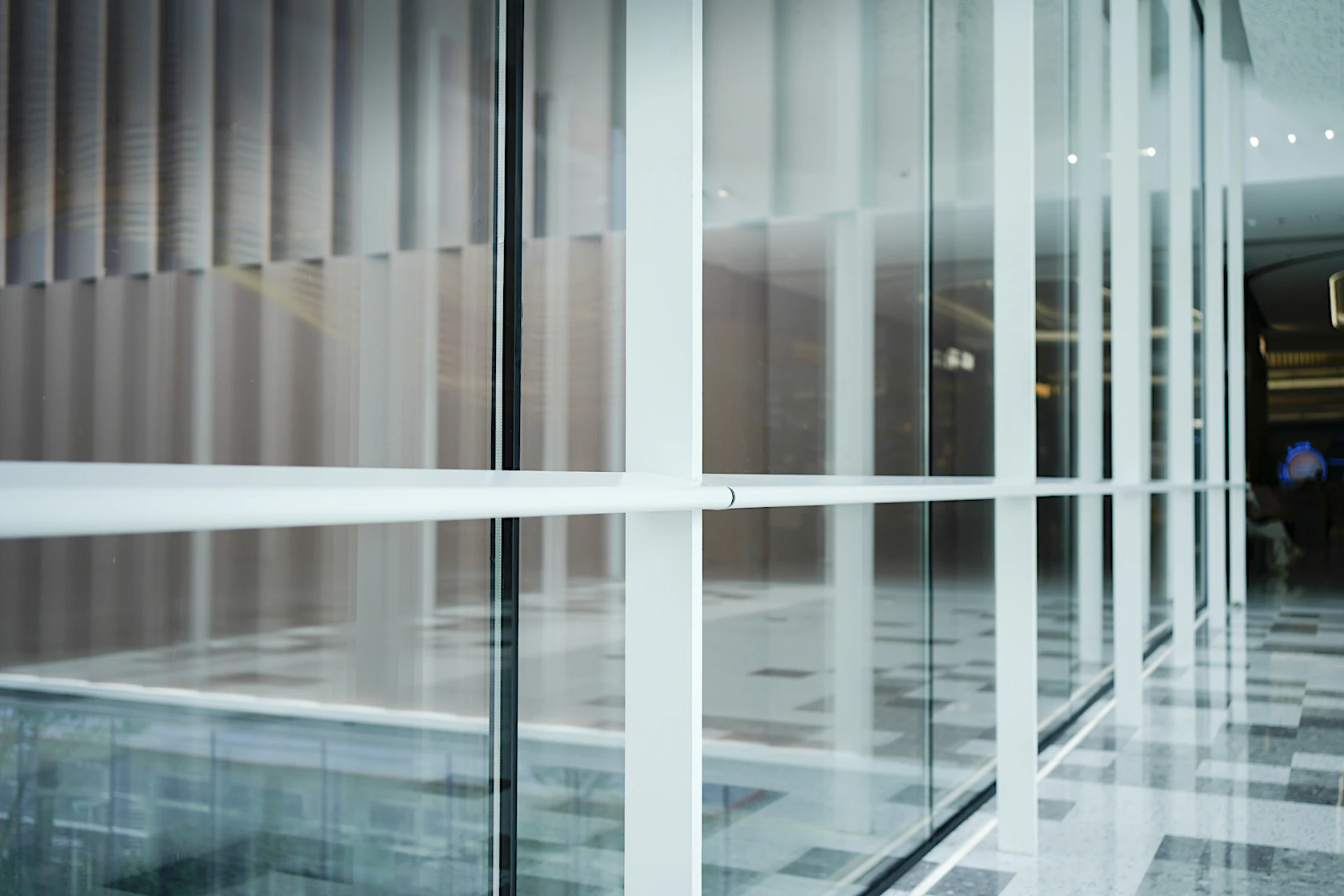 Perspective shot of sleek floor-to-ceiling glass partitions with crisp white metal frames, receding into the depth of a contemporary commercial interior. The semi-transparent glass reveals warm vertical slat panels behind, while the polished checkered stone floor reflects soft ambient light. The strong linear perspective and clean geometric lines create a minimalist, airy aesthetic, typical of modern shopping malls, premium office lobbies or public commercial spaces.